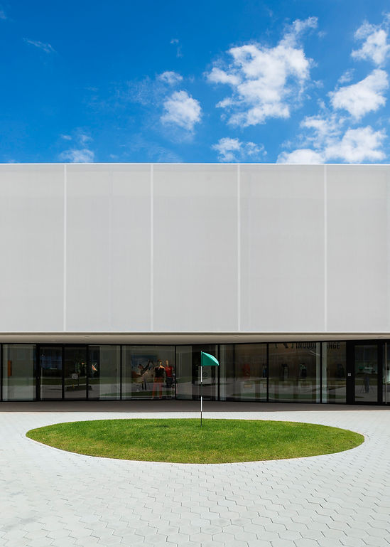 Teilansicht des GolfHouse Bielefeld mit Schaufenstern, darüber eine weisse Stoff-Fassade, im Vordergrund ein gepflasterter Platz mit einer kleinen runden Grünfläche mit Golffahne