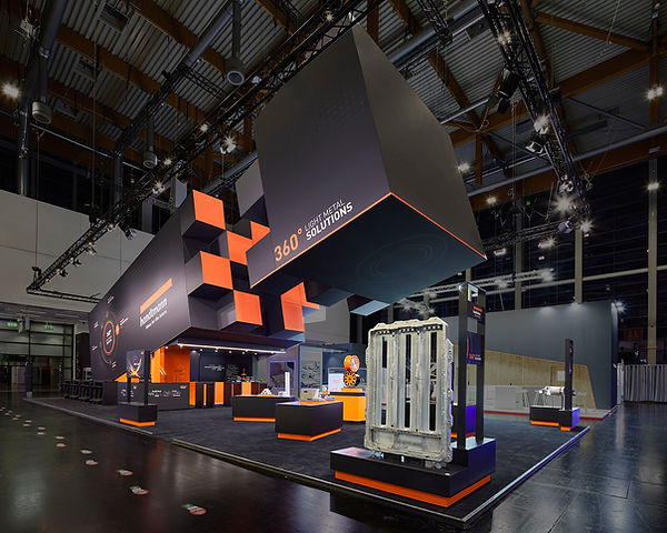 Messestand ohne Besucher in Schwarz und grellem Orange mit einem großen Metallteil an einer Standecke. Unter einem großen, schräg nach oben verlaufenden Aufbau stehen weitere Ausstellungsstücke.