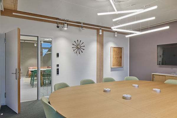 Blick in einen Konferenzraum mit Holztisch und sechs grünen Polsterstühlen, darüber hängt eine moderne Lampe. Die Tür zum Flur steht offen, dort ist ein Stehtisch mit grünen Stühlen zu sehen.
