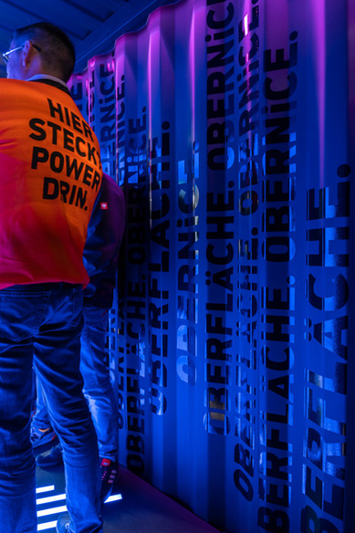 Mann mit orangenem T-Shirt mit Aufdruck "Hier steckt Power drin" steht an einer blau beleuchteten und beschrifteten Wand
