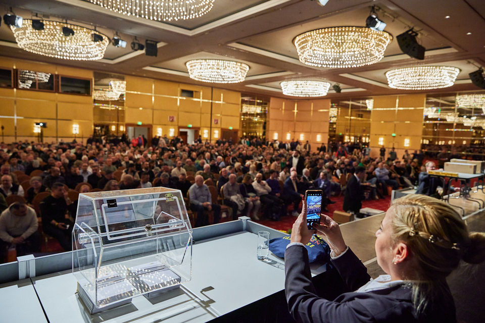 Blick von der Bühne in einen gut gefüllten Kongresssaal, im Vordergrund eine Lostrommel und eine Frau die mit ihrem Smartphone den Saal fotografiert.