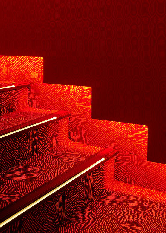 Detail einiger Stufen eines Treppenaufganges. Der Teppich auf den Stufen ist in rotes Licht getaucht.