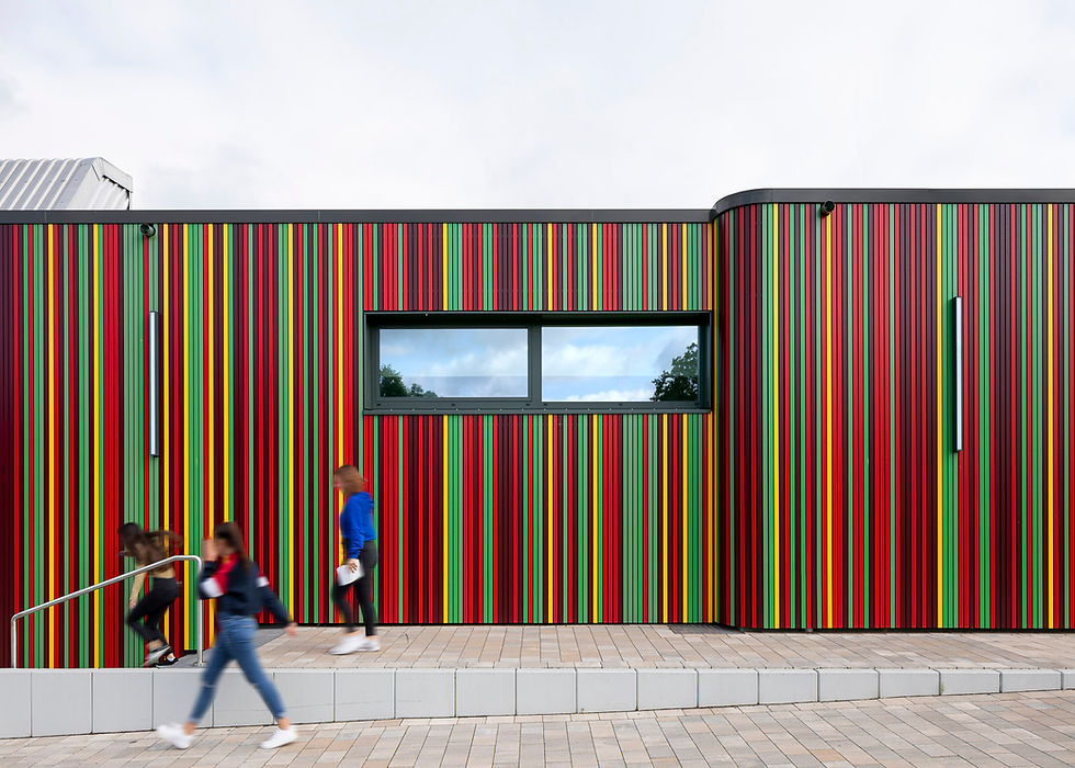 Teilansicht des Schul- und Bürgerforum Barntrup mit einer Fassade aus bunten vertikalen Streifen und einem Fenster, im Vordergrund drei verwischte Schulkinder