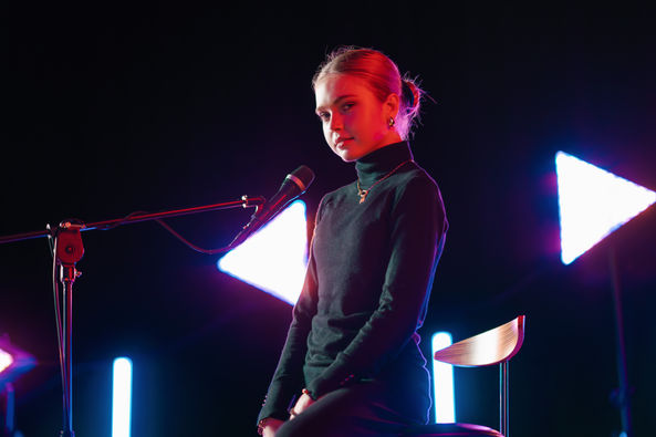 Sängerin sitzt am Mikrofon, trägt schwarzes Oberteil, vor bunten Lichtern. PHOTOGRAPHY