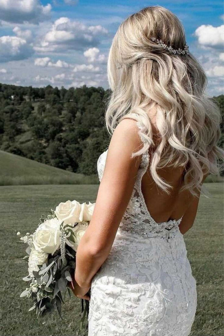 Bride with long blonde hair in a lace gown holds white roses, standing on a grassy hill with a cloudy blue sky backdrop.