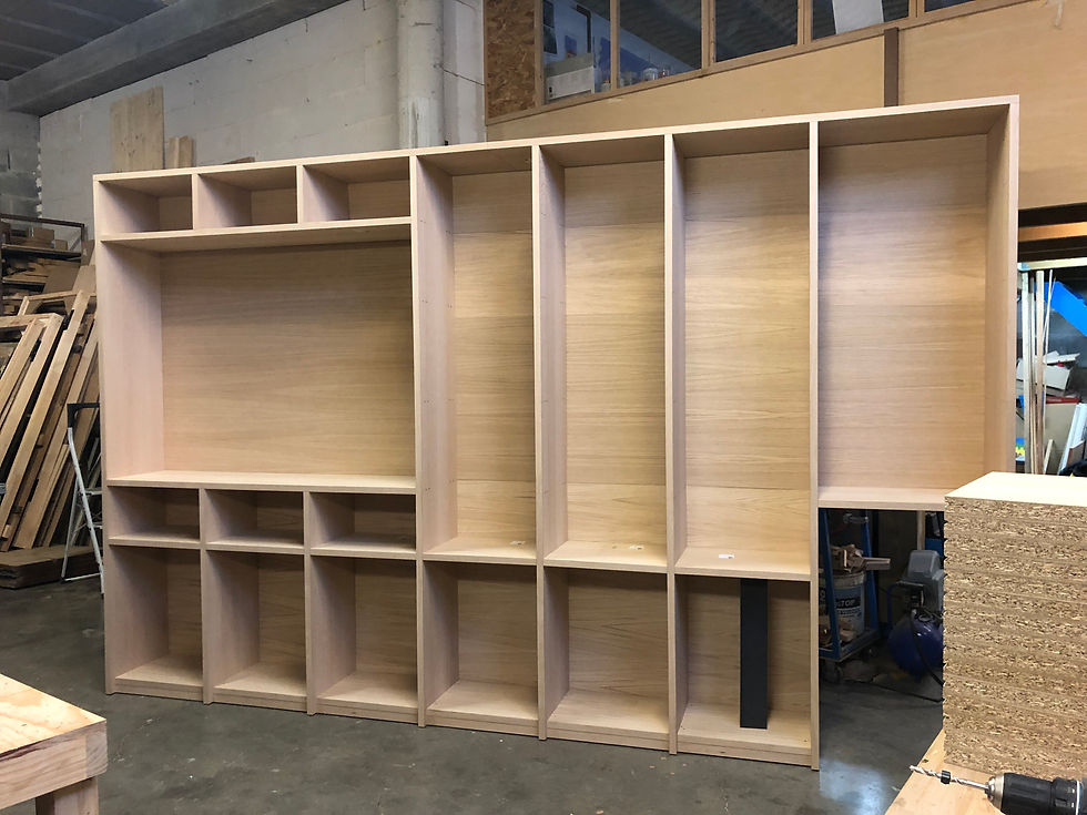 Quentin Delion, cabinetmaker and designer in Bidart, Basque Country — floor-to-ceiling built-in bookcase, alternating open asymmetric niches and closed oak veneer cabinets, set against the wall of a room mid-renovation