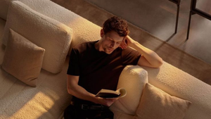 Homem relaxa em sofá bege, lendo um livro. Luz suave e almofadas ao redor criam um ambiente tranquilo e acolhedor.