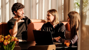 Três pessoas conversam em uma sala iluminada pelo sol, com um laptop na mesa. Tulipas amarelas ao fundo criam uma atmosfera acolhedora.