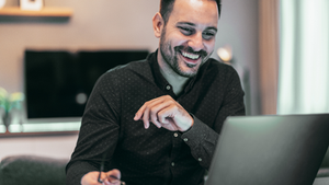 Homem sorridente em roupa escura usa laptop em sala bem iluminada. Ambiente moderno com decoração neutra. Atmosfera de concentração e alegria.