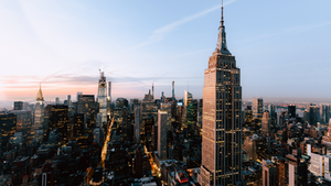 Vista aérea de Nova York ao entardecer. Prédios altos iluminados, céu rosa-azulado. Empire State à direita, atmosfera urbana vibrante.
