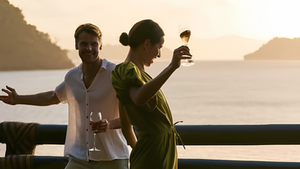 Casal sorri e levanta taças de vinho ao pôr do sol em uma varanda com vista para o mar. Atmosfera alegre e tropical.