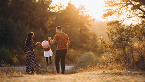 Família em um campo com sol ao fundo. Pais seguram uma criança entre eles; a menina veste chapéu rosa e vestido branco. Atmosfera alegre.