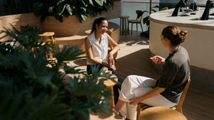 Mulheres conversam em cafeteria iluminada, sentadas ao redor de uma mesa de madeira. Ambiente aconchegante com plantas e bancadas modernas.