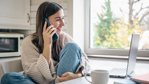 Mulher sorrindo, sentada em cozinha, conversando ao celular. Ela usa suéter bege e segura laptop. Xícara branca sobre a mesa. Ambiente claro.