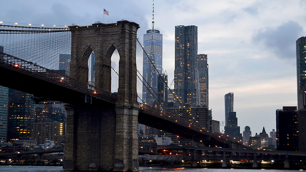 Ponte do Brooklyn ao entardecer, arranha-céus iluminados ao fundo. Céu nublado, bandeira dos EUA no topo da ponte. Atmosfera urbana.