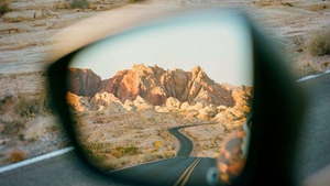 Reflexo no retrovisor mostra estrada sinuosa e montanhas sob sol alaranjado, contrastando com estrada deserta à frente. Atmosfera tranquila.