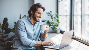 Homem sorridente toma café enquanto usa laptop perto de janela. Plantas no fundo; atmosfera leve e descontraída.
