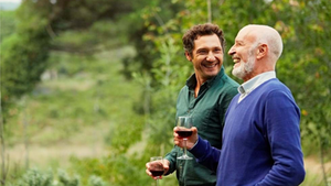 Dois homens sorrindo e conversando ao ar livre, segurando taças de vinho. Ao fundo, vegetação verde. Atmosfera alegre e amigável.