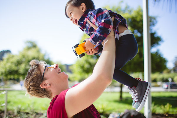 White woman with light brown hair and glasses is smiling holding up a small child who has a toy truck in his hands. Child is about 2 and smiling.