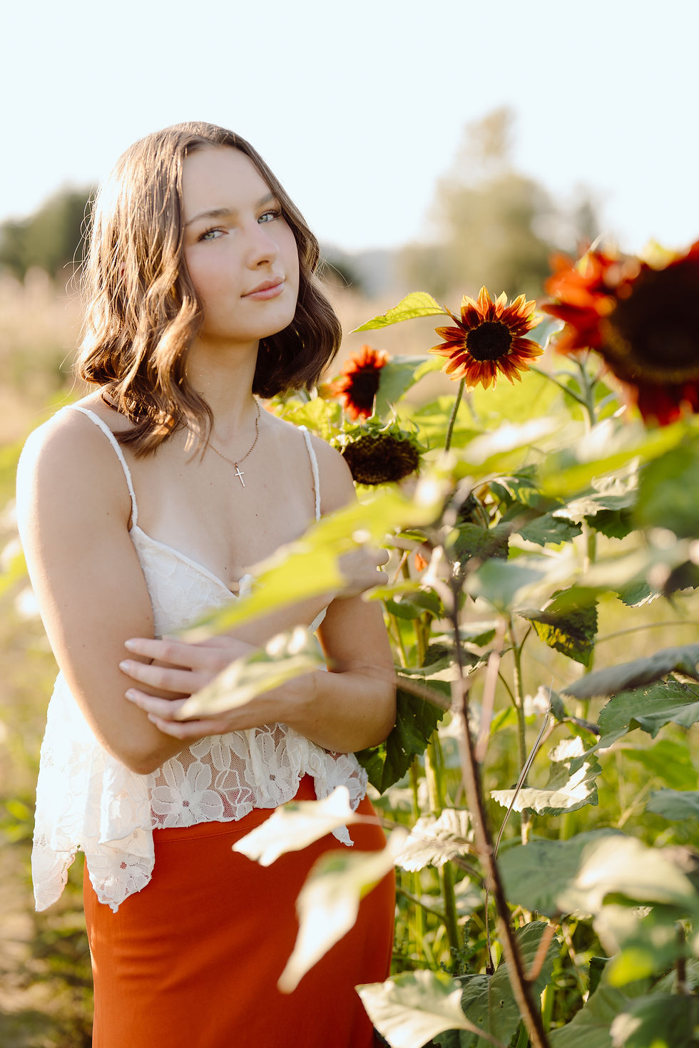 Woman in white lace top and orange skirt stands in sunflower field, arms crossed, with a serene expression. Sunlight and greenery surround.