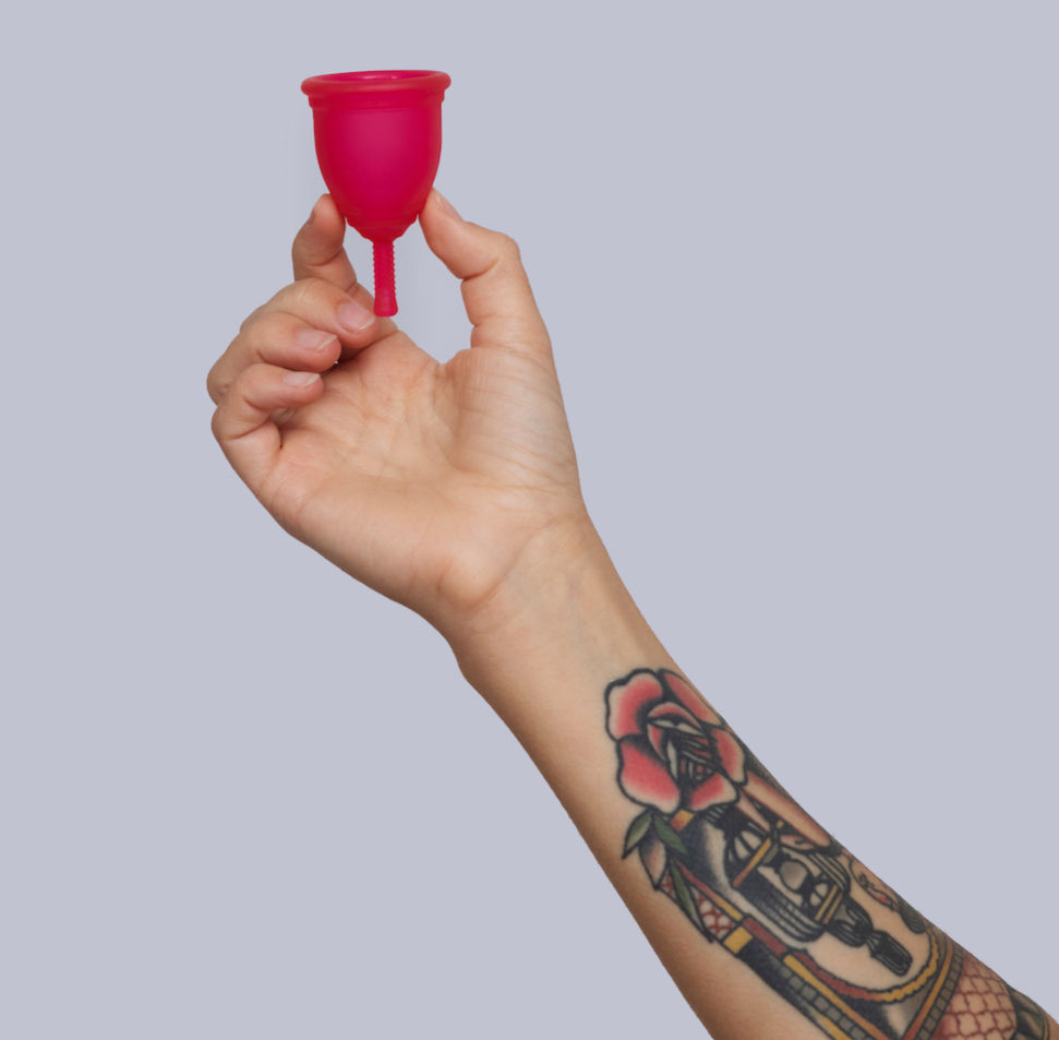 A ladies hand with a tattoo on her arm, holding up a pink menstrual cup from Ruby Cup.