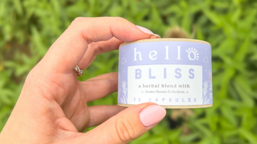 Hand holds a herbal supplement pot labeled "hello BLISS" with 30 capsules of herbal blend. Greenery in background, mood is calm and natural.