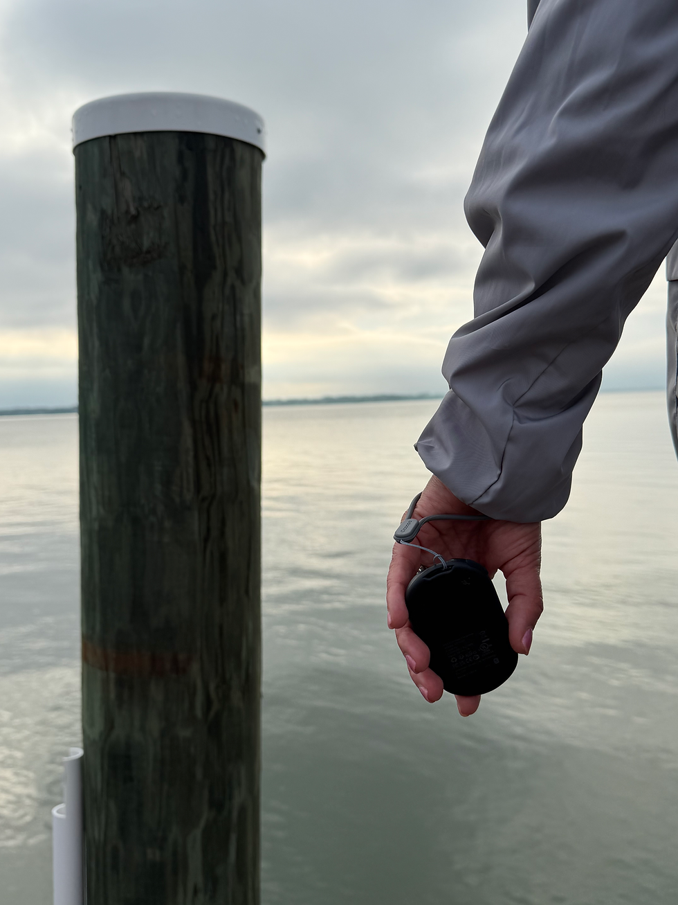 Person in a gray sleeve holds a black hand warmer beside a weathered dock post over calm water under a cloudy sky.