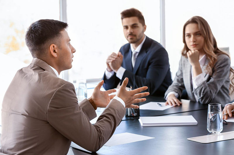 Mann sitzt gestikulierend gegenüber eines Mannes und einer Frau am Tisch