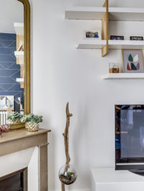 décoration de salon, cheminée ancienne, miroir doré, miroir Louis Philippe, étagères murales blanches, bois et blanc, meuble TV, salon contemporain, papier peint géométrique, mur bleu nuit, objets décoratifs, plantes succulentes, terrarium suspendu, cadre photo, aménagement salon, style scandinave, décoration murale, intérieur moderne, appartement parisien, design épuré, salon chic.