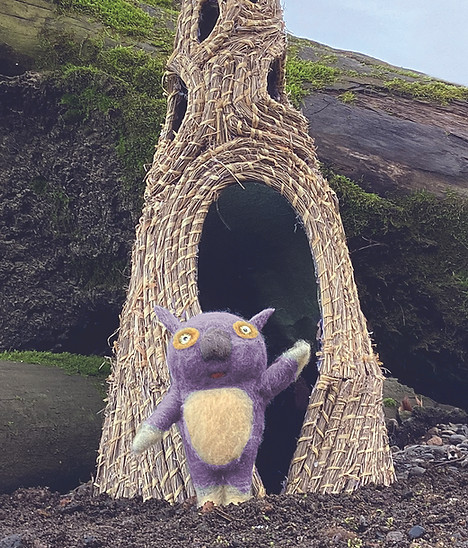 Purple critter waving in front of a tall woven sculpture home. Created by Catherine Failor