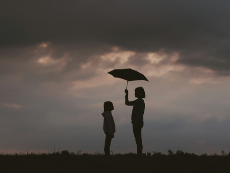 Silhouette einer erwachsenen Person, die einem Kind mit einem Regenschirm Schutz bietet vor einem dunklen Himmel. Symbolbild für elterliche Fürsorge und mögliche Überbehütung.
