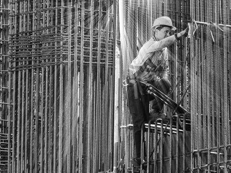 Bauarbeiter mit Schutzhelm arbeitet zwischen hohen Stahlarmierungen auf einer Baustelle und überprüft eine Konstruktion im Rahmen sicherer Arbeitsabläufe.