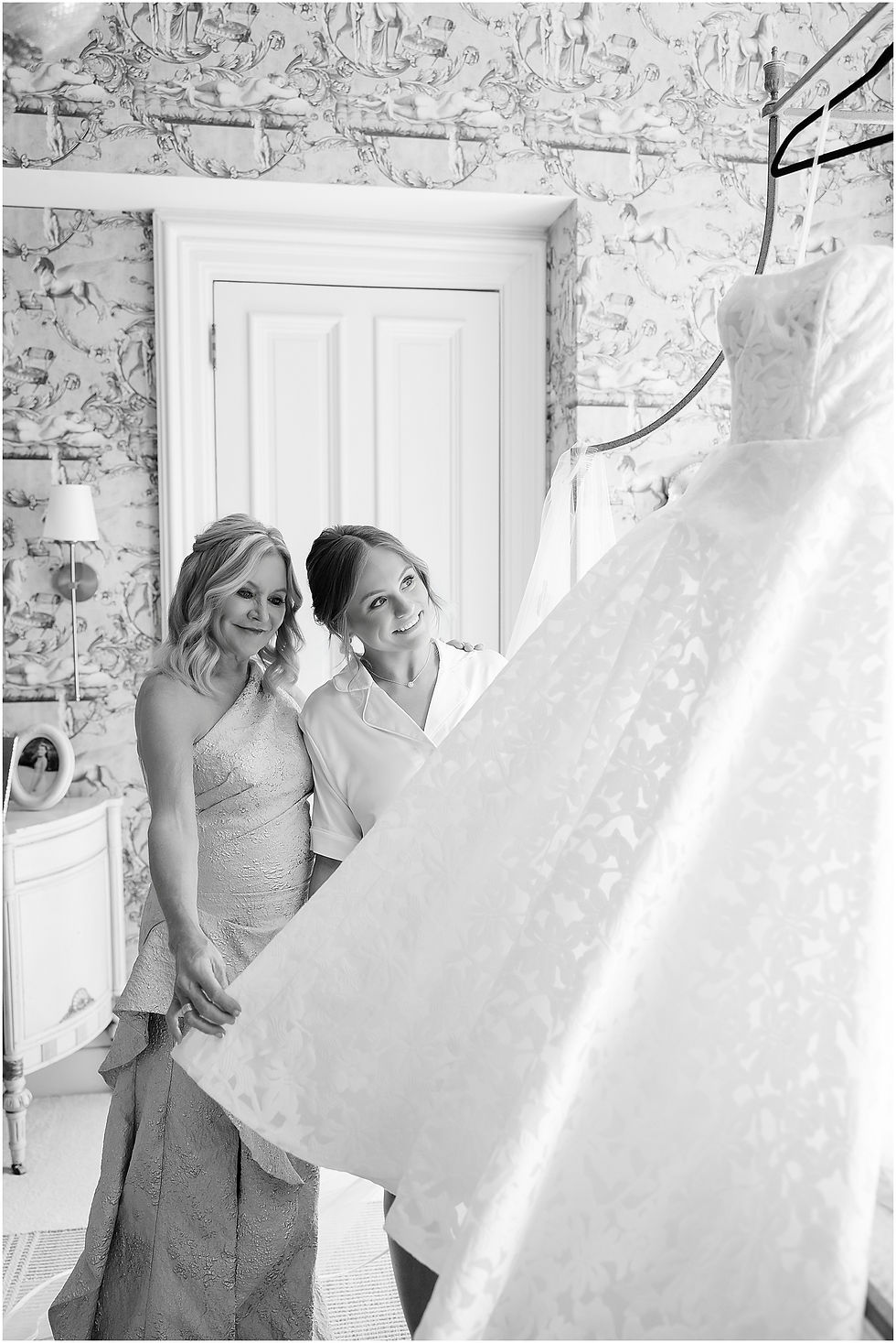 Two women smile at a hanging wedding dress in a black-and-white bedroom with horse-pattern wallpaper.