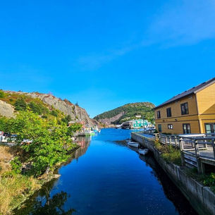 Calm blue river reflecting the sky, flanked by trees and colorful buildings. Rugged hills in the background under a clear, bright sky.