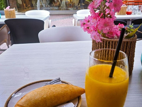 A table with a pastry on newspaper, orange juice, and pink flowers. Background has shop mannequins. Bright, casual setting.