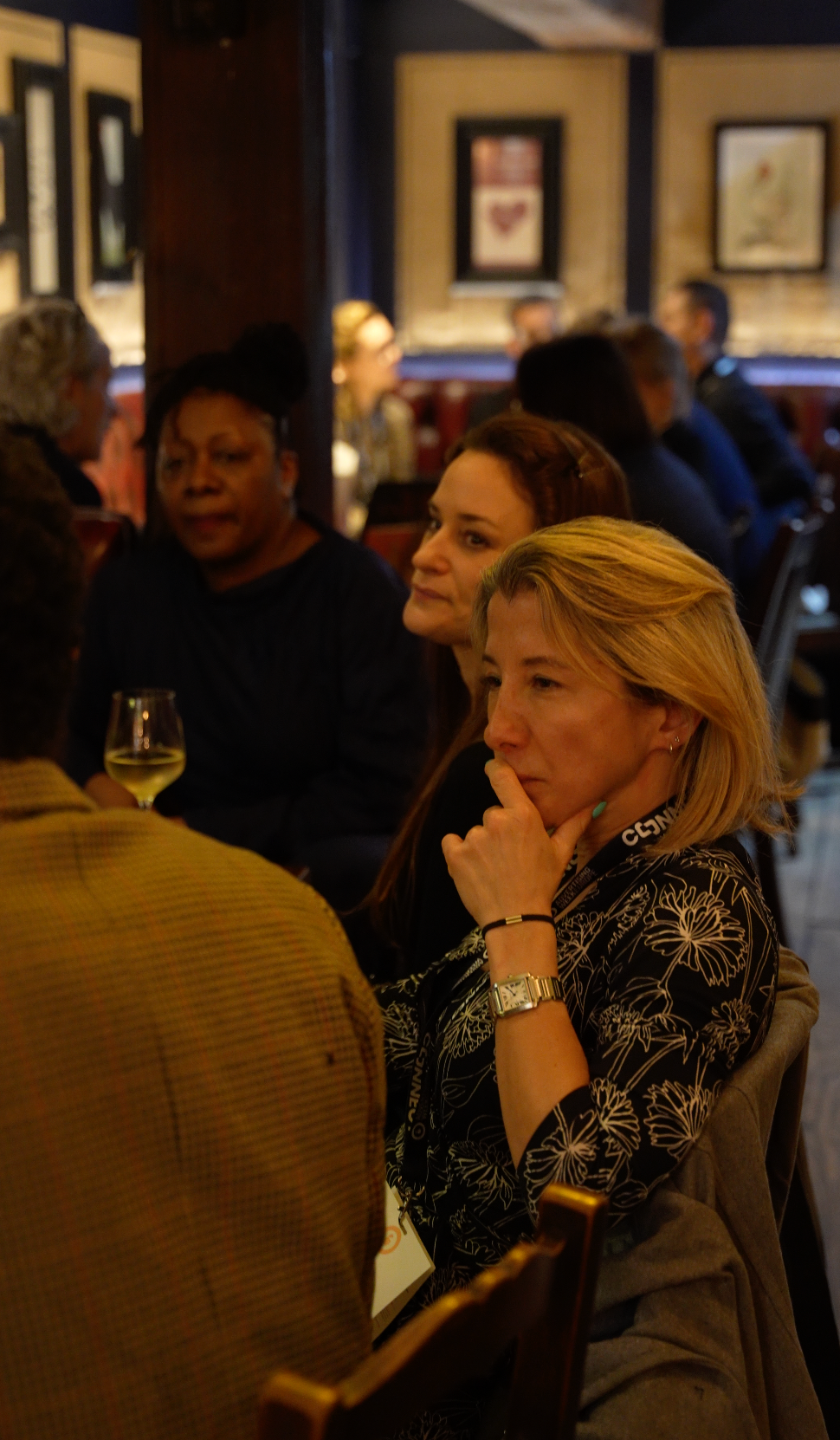 Three people sitting at a dimly lit restaurant table, engaged in conversation. One woman, deep in thought, rests her chin on her hand.