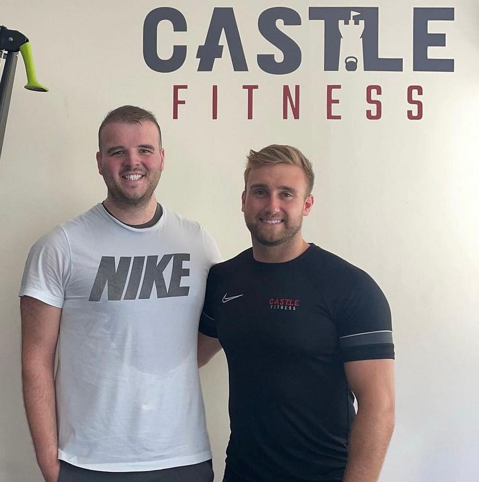 Jake and Brad are smiling in front of the Castle Fitness logo at the studio. Jake is wearing a white Nike top and Brad is wearing a black top.