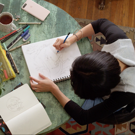 A woman as seen from above, drawing on a sketchbook while sitting at a table. 