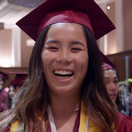 A high schooler wearing a maroon graduation cap and coat smiles. 