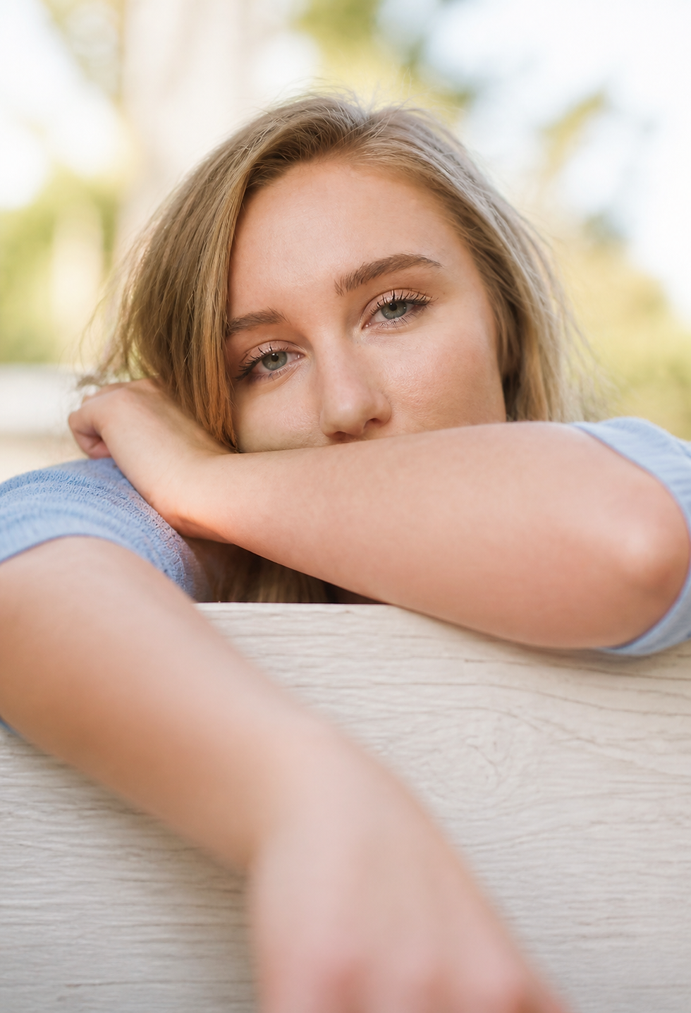 Jeune femme aux cheveux blonds, appuyée sur une clôture blanche, regard calme en extérieur, fond flou lumineux. Maman d'Amour.