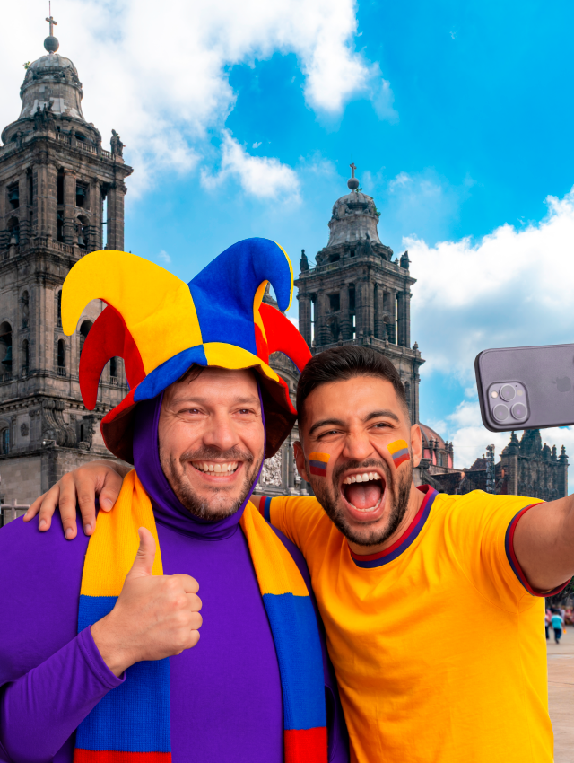 Selfie de dos hinchas de Colombia: uno con gorro tricolor y pulgar arriba, el otro sonriendo, con la catedral mexicana detrás.