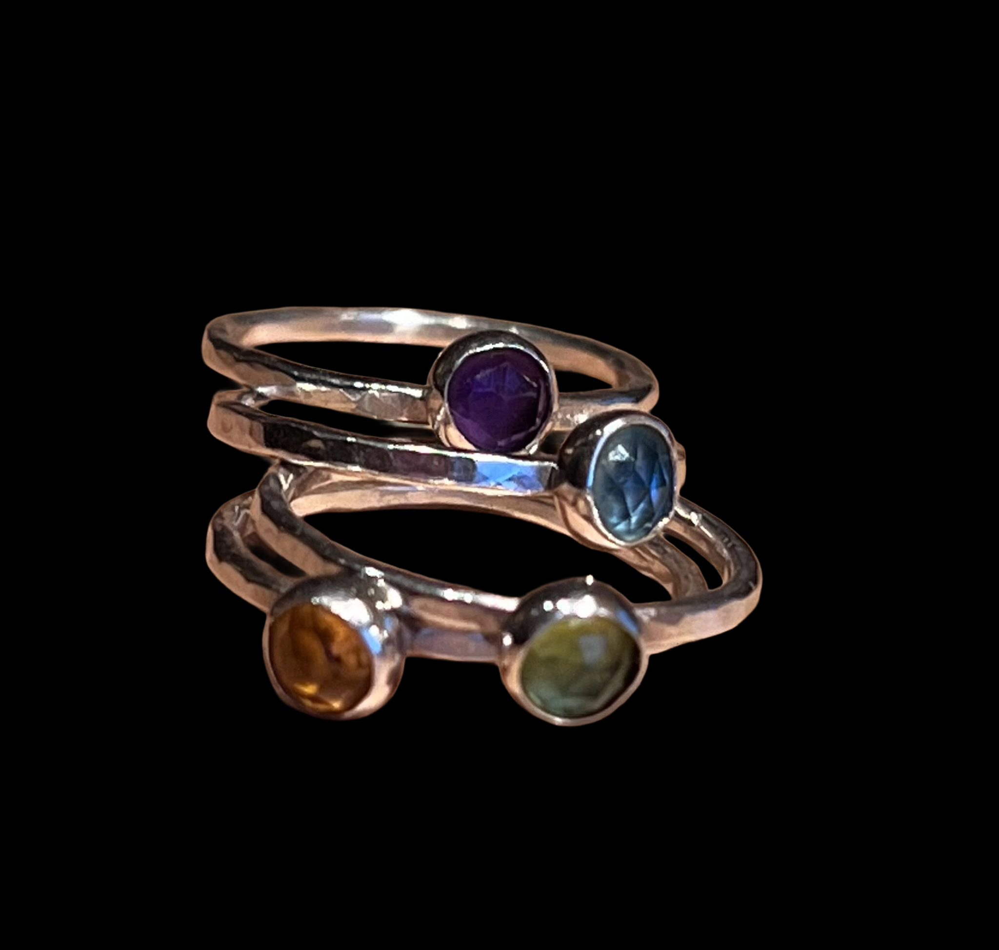 Close-up product photo of four handcrafted sterling silver stacking rings arranged together against a black background. Each ring features a bezel-set faceted gemstone in a different color: deep purple, sapphire blue, warm amber, and soft green. The rings have lightly hammered bands and a polished finish, highlighting the handmade craftsmanship and colorful gemstone accents.