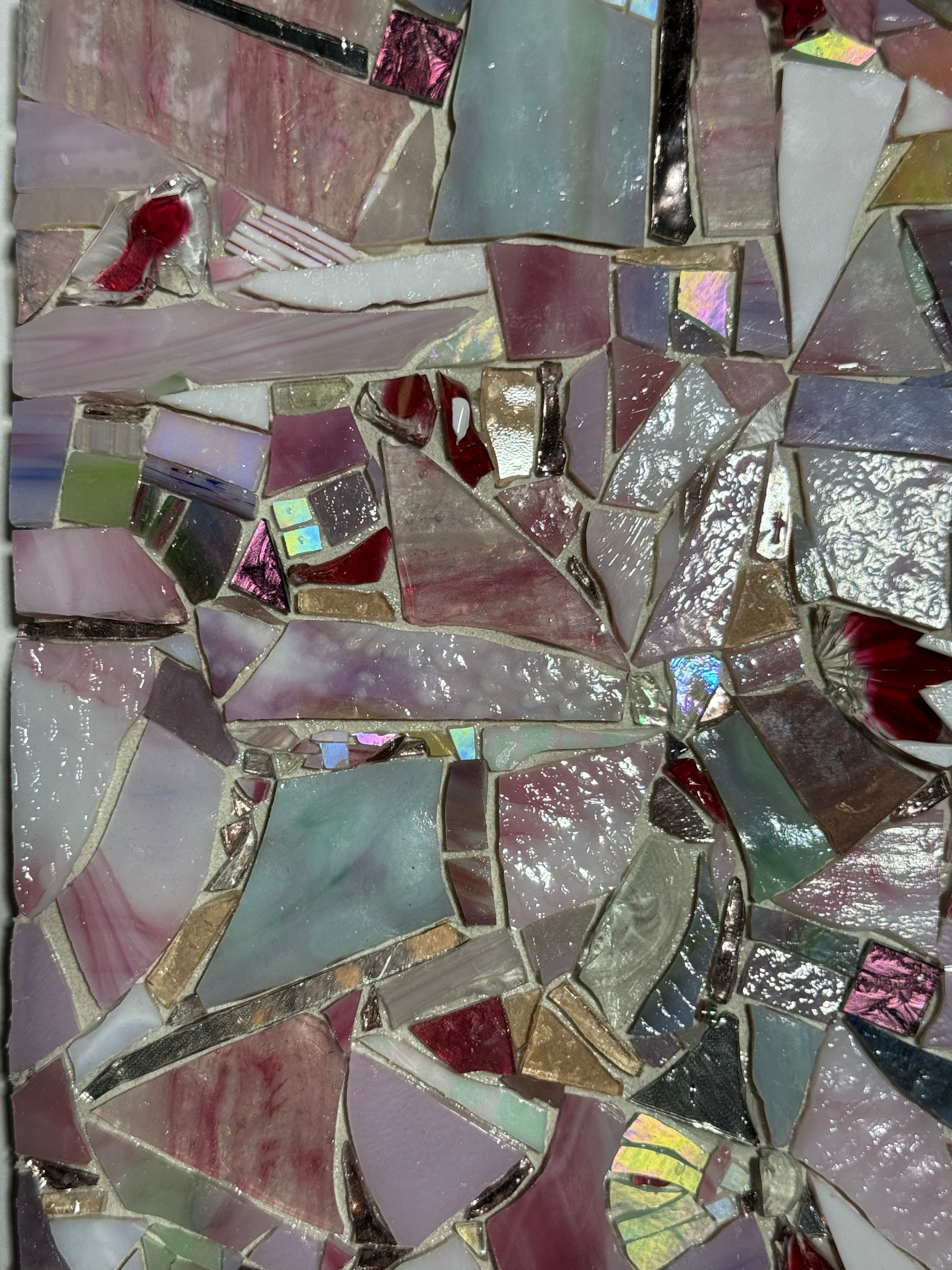 A close-up view of handcrafted mosaic tesserae arranged in an intricate composition. Varied shapes, textures, and reflective surfaces of glass, stone, and mixed materials create movement, depth, and rich visual detail throughout the artwork.