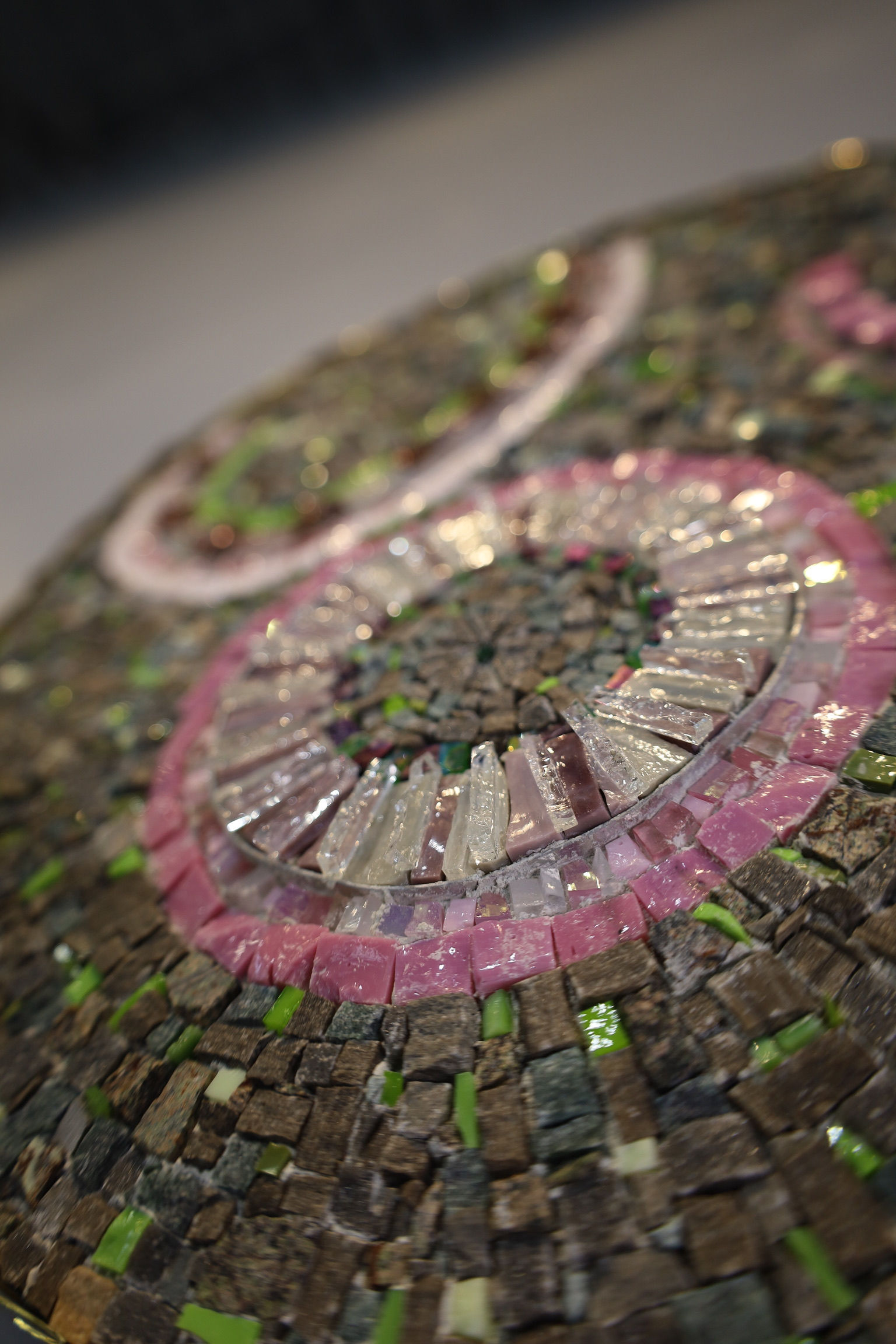 A close-up view of handcrafted mosaic tesserae arranged in an intricate composition. Varied shapes, textures, and reflective surfaces of glass, stone, and mixed materials create movement, depth, and rich visual detail throughout the artwork.