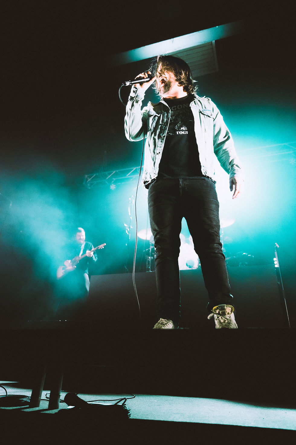 Singer on stage with microphone, vibrant teal lights in the background, wearing a denim jacket. Another musician plays guitar, creating an energetic mood.