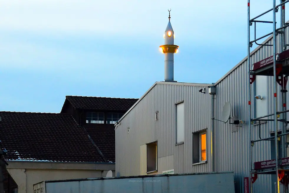 Mosque in Switzerland with a lit minaret rising above industrial-style buildings at dusk.