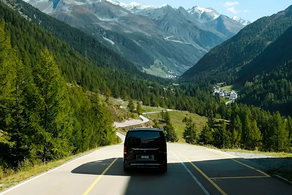 A black Noir van drives along a winding alpine road between green valleys and snow-capped peaks, illustrating how a swiss chauffeur service covers every mile of your journey in comfort.