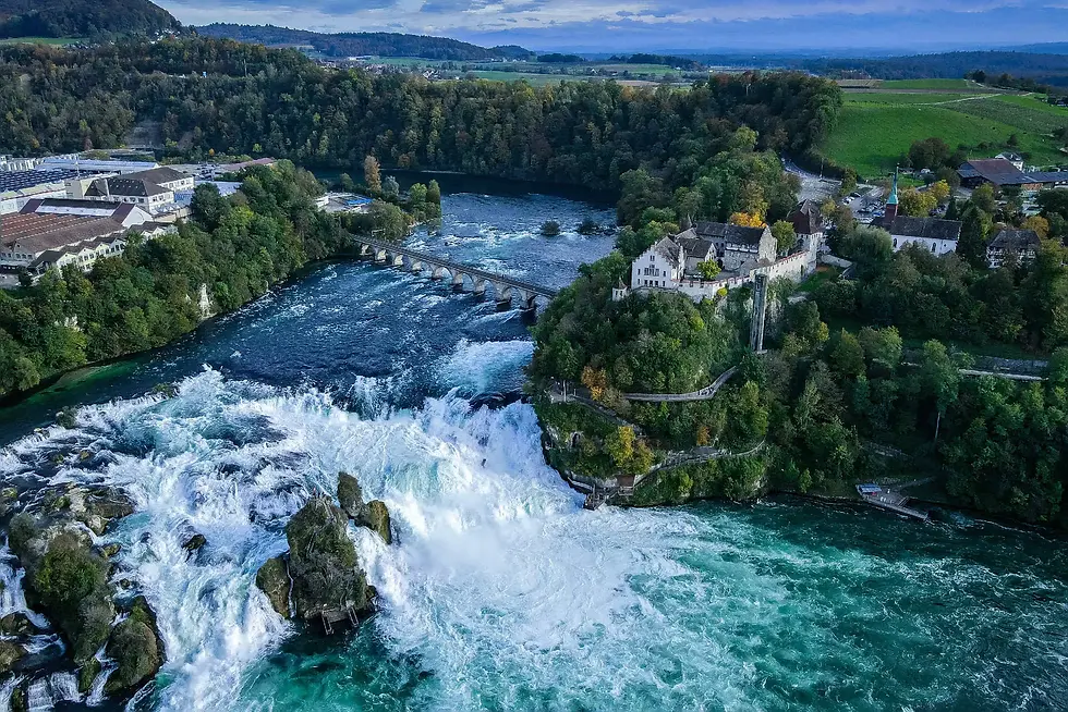Foto udara Rhine Falls Switzerland in winter menampilkan air terjun besar yang mengalir deras di antara tebing hijau, jembatan batu, dan kastel tua di puncak bukit.
