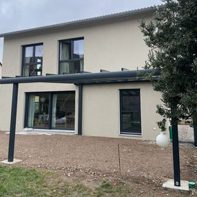 Montagefortschritt einer Terrassenüberdachung an einem modernen Wohnhaus; bereits installierte Aluminiumkonstruktion mit Pfosten und Querträgern, offener Bodenbereich ohne Belag, große Fenster und Schiebetüren an der Fassade, seitlich ein Baum und Gartenfläche – Dokumentation eines Bauabschnitts für Überdachungssysteme von ML DER RAUM.