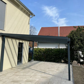 Fertig montierte Terrassenüberdachung mit schlanker Aluminiumkonstruktion in dunkler Farbe an einem modernen Wohnhaus; darunter großformatige, helle Terrassenfliesen, daneben eine dichte Gartenhecke und ein Nachbargebäude mit rotem Ziegeldach, klarer Himmel im Hintergrund – Abschlussdokumentation eines Überdachungsprojekts von ML DER RAUM.